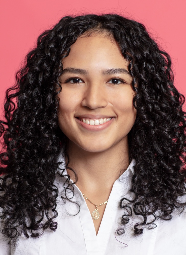 A person with curly black hair, wearing a white shirt and a gold necklace, smiles in front of a pink background.